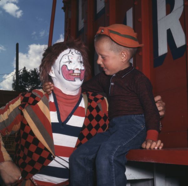 A man dressed as a clown is standing next to a young boy at Circus World Museum. The boy is sitting on the side of a circus wagon.