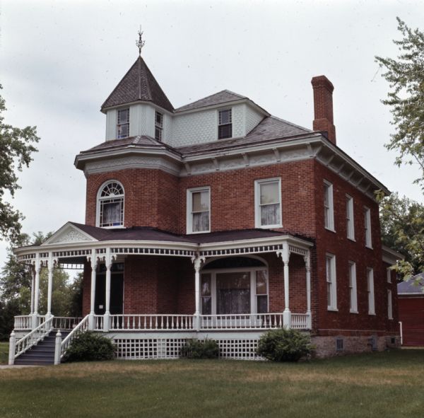 View across front lawn towards the historic Beyer Home. The front porch wraps around the side of the house to the left, there is a three-story tower room and entry, and some of the windows have leaded glass.