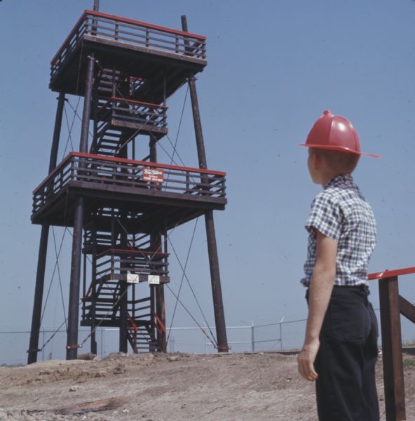 A young boy wearing a red plastic firefighter hat is looking up at a five-story fire lookout tower at the Hall of Flame Fire Museum.