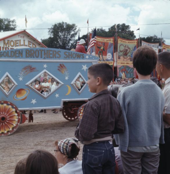 View from rear of a crowd of people watching circus wagons passing by on a dirt road at Circus World Museum. Two horses are pulling a blue wagon with red and orange wheels with the words "Golden Brothers Shows" painted at the top of the wagon. Large banners advertising circus acts are hanging in the background.