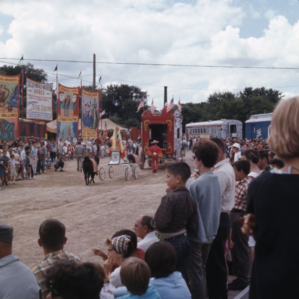 View from a crowd of people watching horse-drawn circus wagons parading on a dirt road at Circus World Museum. Behind the wagon is a man dressed in a red and orange costume leading a lama pulling a cart. A woman in a matching costume and headdress is riding in the cart, and another lama is following behind. Banners advertising circus acts are hanging on the opposite side of the road.
