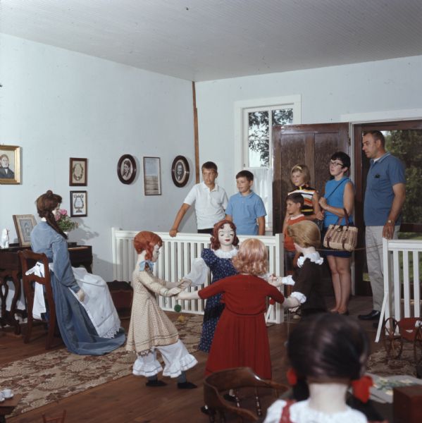 View from back of display of people visiting the first kindergarten building. A mannequin dressed as a nineteenth century teacher is sitting in a chair on the left, and four mannequins dressed as girls are holding hands and standing in a circle. Pictures are hanging on the wall, including a portrait of Margarethe Schurz and her husband Carl Schurz.
