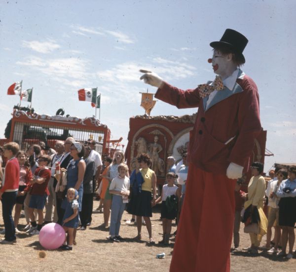 A clown on stilts is pointing towards the left. Behind him a crowd of people are standing in a line. Two circus wagons are behind the crowd, one an animal cage flying the Mexican flag, the other decorated with gold relief designs of a woman riding a lion.