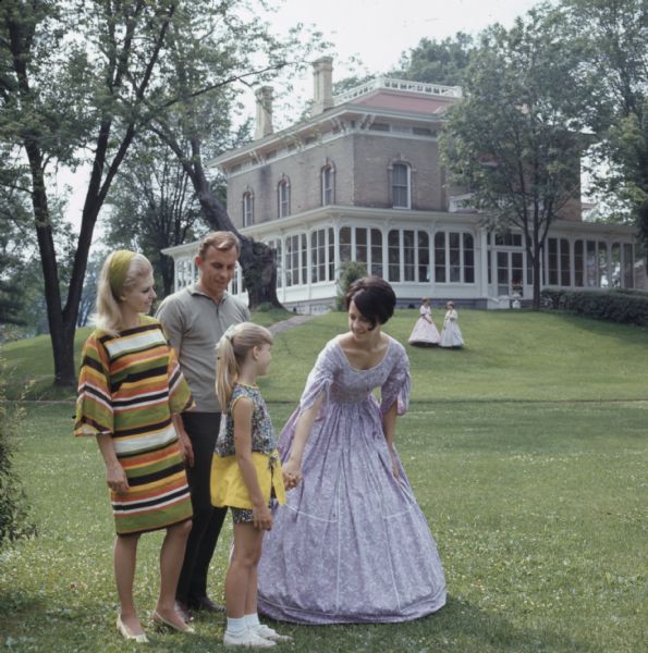 A woman dressed in period clothing is holding the hand of a young girl, with her parents standing behind her. Two other women in period clothing are standing on a hill in front of Villa Louis in the background.
