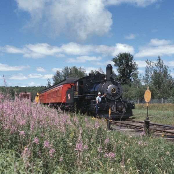 View from side of railroad tracks towards a man posing at the front of a vintage steam locomotive. Flowers are in the foreground.