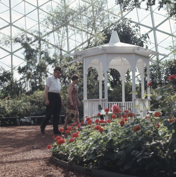 A man and woman are walking along a dirt path near a gazebo. Flowers and trees and bushes are growing inside the horticultural conservatory.