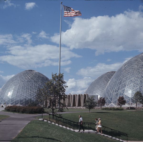 View towards the three glass domes at the Mitchell Park Conservatory. In the foreground three people are walking up a stairway on the grounds of the park. The American flag is flying a tall flagpole near the entrance to the conservatory.