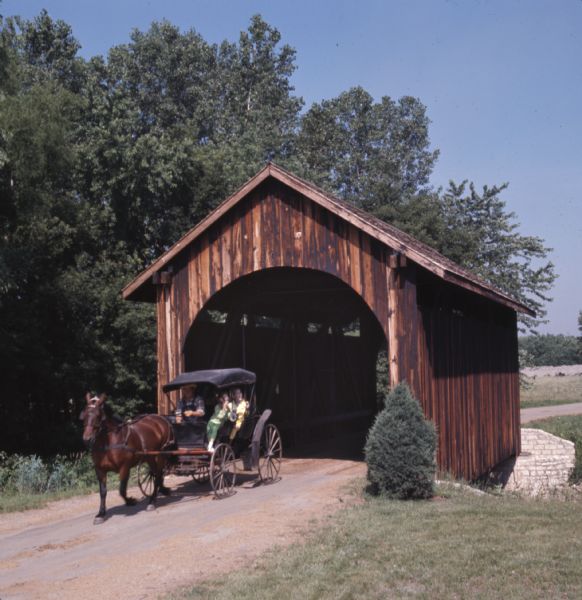 A horse-drawn carriage carrying visitors is coming through a covered bridge at Stonefield Village.
