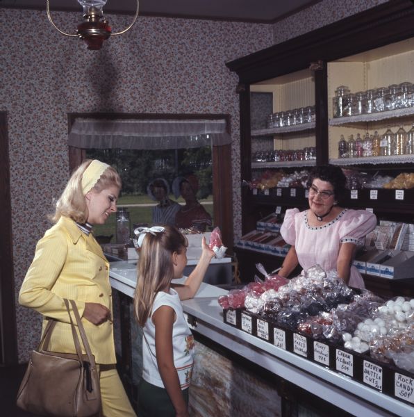 A young girl is holding up a bag of rock candy at the counter of a candy store in Stonefield Village. Her mother is standing behind her. A woman, dressed in period clothing, is behind the counter. Candy is displayed in boxes and jars along the counter and on shelves in the background. A painting of two women in period dress, made to look like a scene through a window, is hanging on the wall.