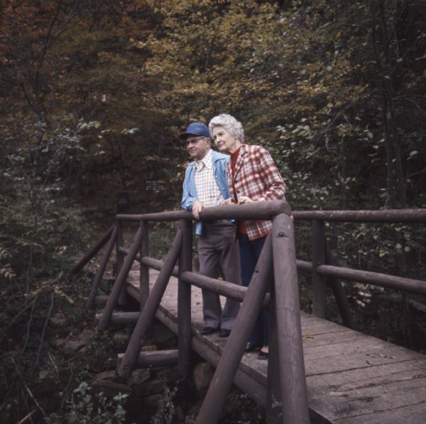 Melvin and Sadie Tvedt, of Mount Horeb, standing at the railing looking towards the left on a wooden bridge in Governor Dodge State Park. The trees around them are displaying fall colors of orange and red.