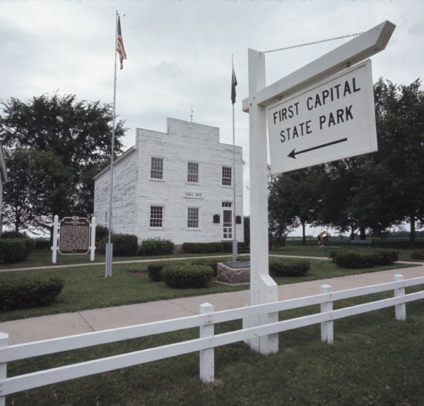 A white sign in the foreground near a fence reads: "First Capital State Park," with an arrow pointing towards a white building in the background that has a sign above a window that reads: "Council House." A historic marker is in front of the building on the left. In the center, surrounded by a sidewalk, are two flags on flagpoles, the American and the Wisconsin flag, and an historic marker plaque.