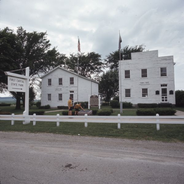View across road towards a white sign that reads: "First Capital State Park" that includes an arrow pointing towards two white buildings in the background, the Council House and the Supreme Court. A historic marker is between the two buildings. In the center is an American and Wisconsin flag on flagpoles flanking two boys looking at a plaque on a stone.