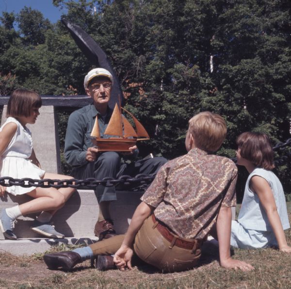Three children sitting outdoors around an elderly man. The man is wearing a ship captains hat and is holding a wooden model of a ship in his lap. The group is sitting around the stone pedestal displaying a large anchor. 