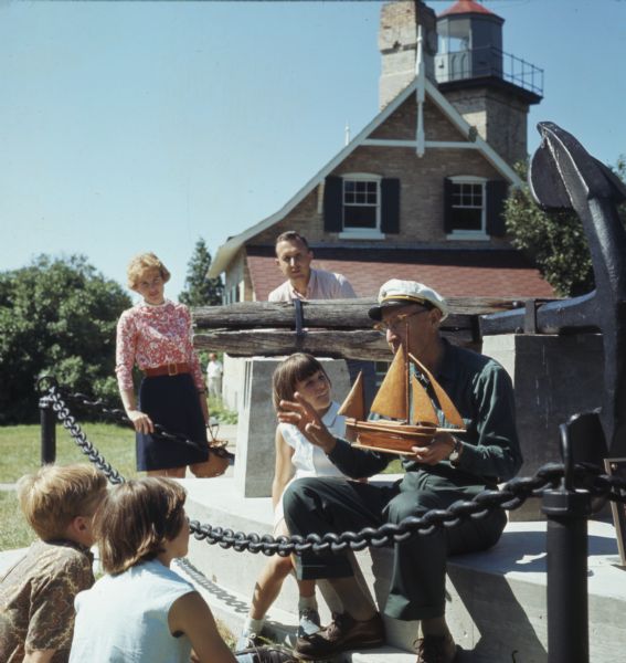 Three children and two adults are stand and sitting around an elderly man wearing a ship captains hat. He is holding a wooden model of a ship. The group of people is gathered around stone pedestal supporting a large anchor. In the background is the Eagle Bluff Lighthouse.