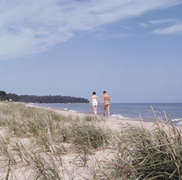 View across grass and sand towards a young couple, a man and woman, holding hands and walking along the shoreline of Whitefish Bay. Trees are along the curving shoreline in the background.