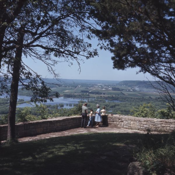 Slightly elevated view of a family standing in front of a stone wall at Point Lookout in Wyalusing State Park. The man is pointing towards the Wisconsin River and Mississippi River below them. The southern portion of Prairie du Chien is in the distance.