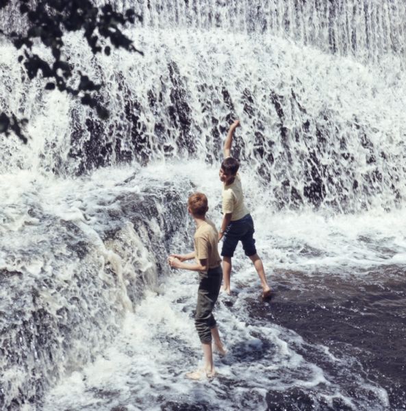 Two boys are playing in the water at the base of a waterfall.