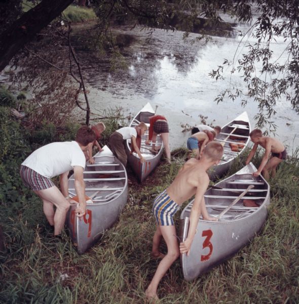 A group of young boys are pushing four canoes down a grassy incline into a lake.