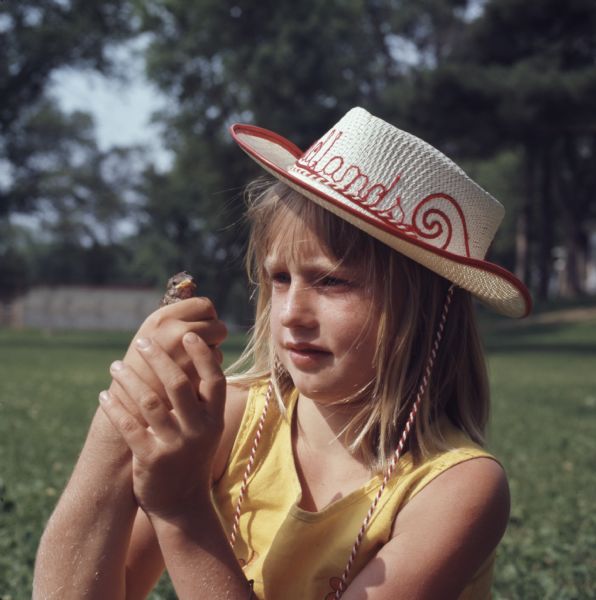 A young girl is holding a baby bird in her hands. She is wearing a red and white straw cowboy hat.
