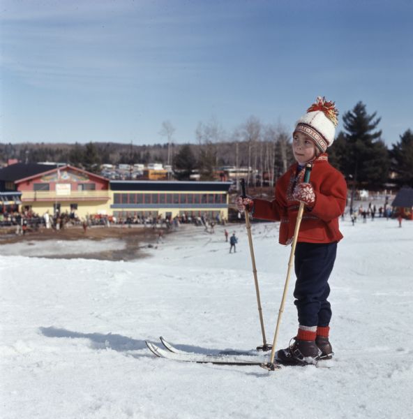 A young child, wearing a Scandinavian style hat, coat, and gloves, is wearing a pair of skis and standing on a small hill. Below the hill in the background is a crowd of people standing outside of the Telemark Lodge. 