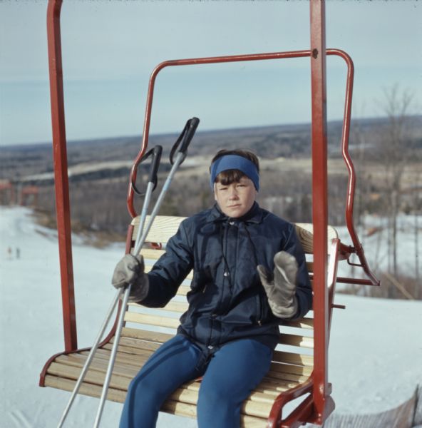 A young boy is sitting on a wood and metal ski lift holding a pair of ski poles and waving to the photographer.