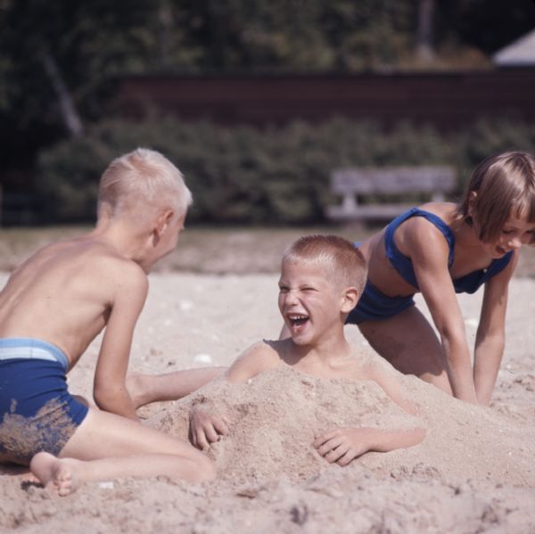 Two children, a boy and girl, are burying another boy in the sand.