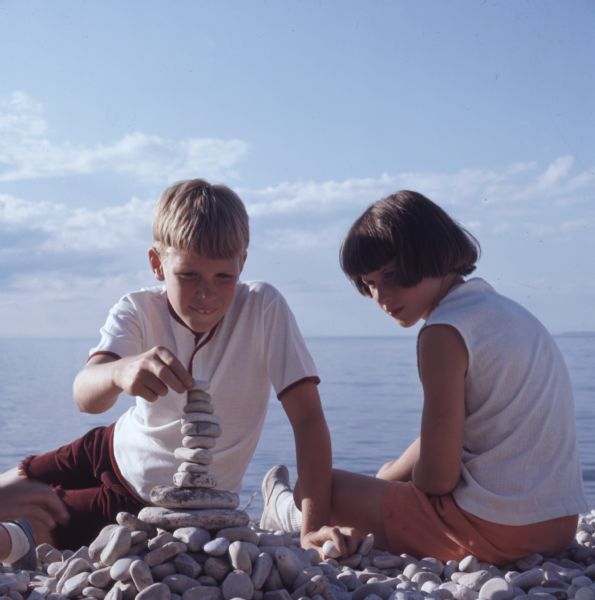 Two children sit on a shingle beach with Lake Michigan in the background. The girl is watching as the boy is stacking stones on top of each other to make a cairn.