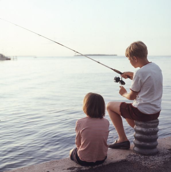 A young boy and girl are sitting on a pier looking out towards the lake. The boy is sitting on a post fishing with a fishing rod.
