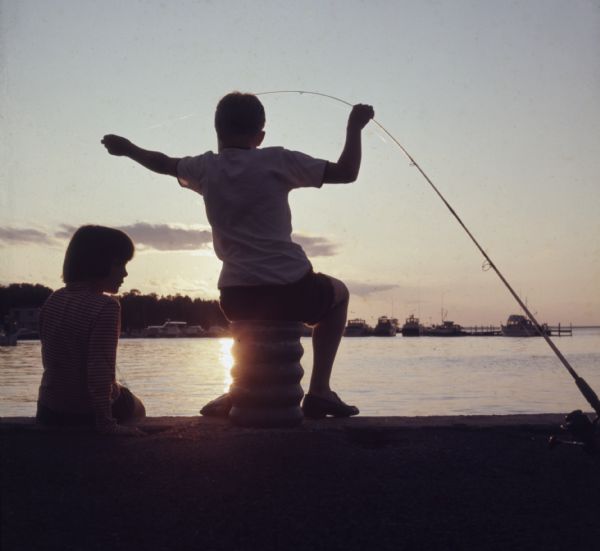Two children are sitting on a pier, silhouetted by the setting sun. The boy is holding a fishing pole in one hand, and pulling the fishing line in another hand. Boats are docked at a pier across the water.