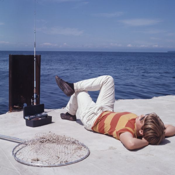 A young boy is lying on his back with one leg crossed over his knee on a concrete pier on a lake. A fishing net and a tackle box is next to him, and a fishing pole is leaning against a metal post.