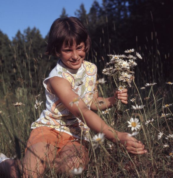 A young girl is sitting in tall grass picking daisies.
