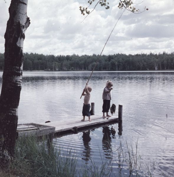 View from shore of two young children standing on a small wooden dock. The boy is holding up a cane fishing pole, and the girl is holding up a stringer of fish.