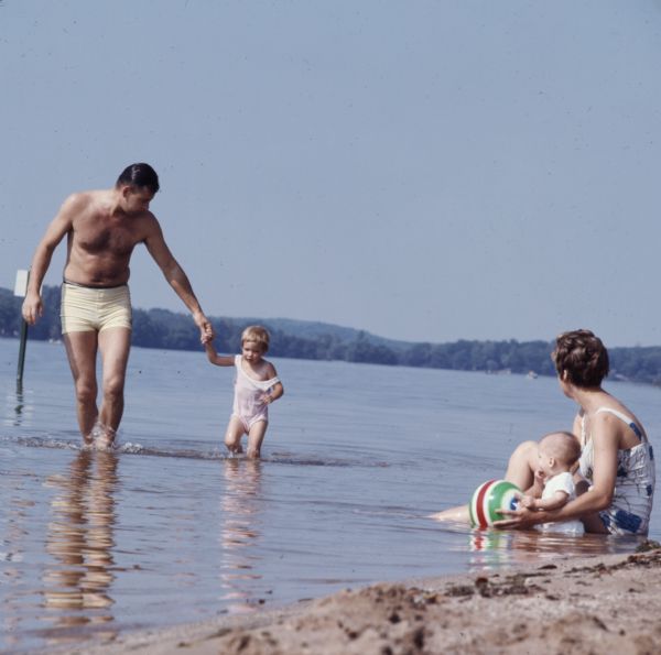 A woman is sitting with a baby and holding a beach ball while sitting in the shallow water on a sandy shoreline. Nearby a man and child are holding hands and walking in the water. There is a sign posted in the water in the background.