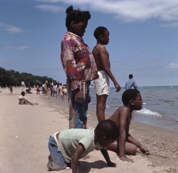 A group of African American children are playing in the sand on Bradford Beach. Other children and adults are wading into the lake or are sitting on the sand behind them.