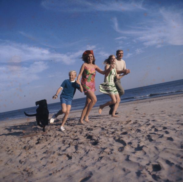 A family of four are running along a beach holding hands, all smiling at the photographer. The young boy is holding the leash of a black lab.