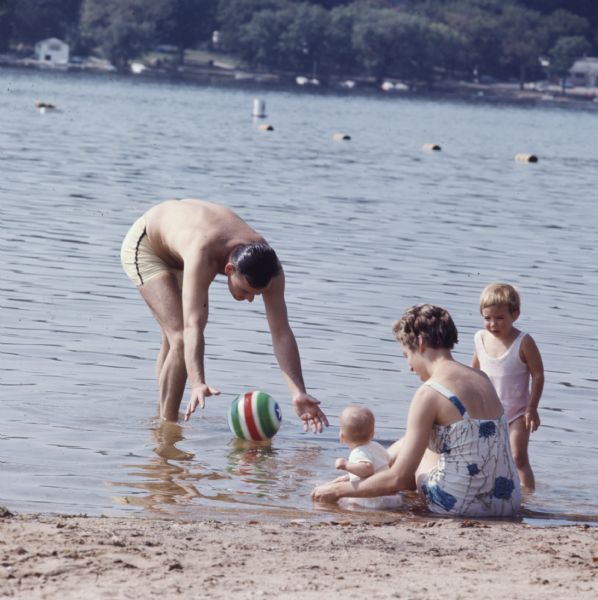A family is playing in the water at a beach. The mother is sitting in the shallow water at the shoreline, holding her baby. Another child is standing next to her. The father is leaning down over a beach ball floating in the water.