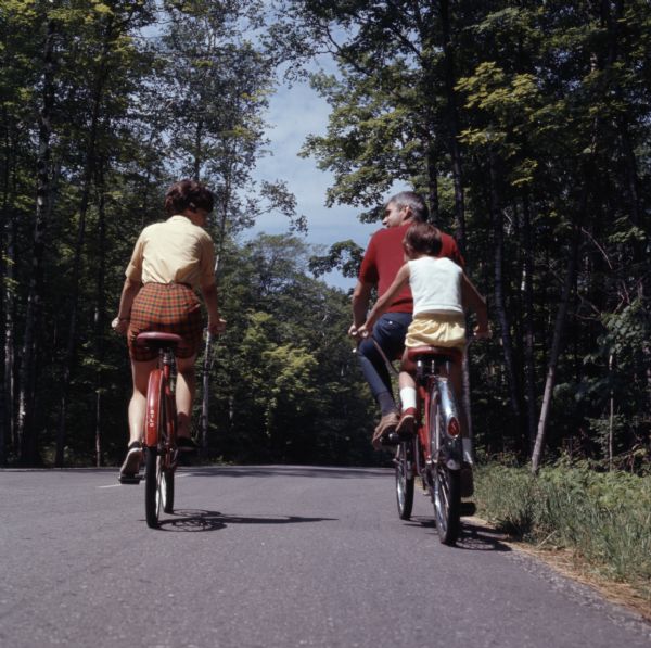 View from behind of a family of three biking down a paved road in Peninsula State Park. The child is riding a tandem bicycle with the father.