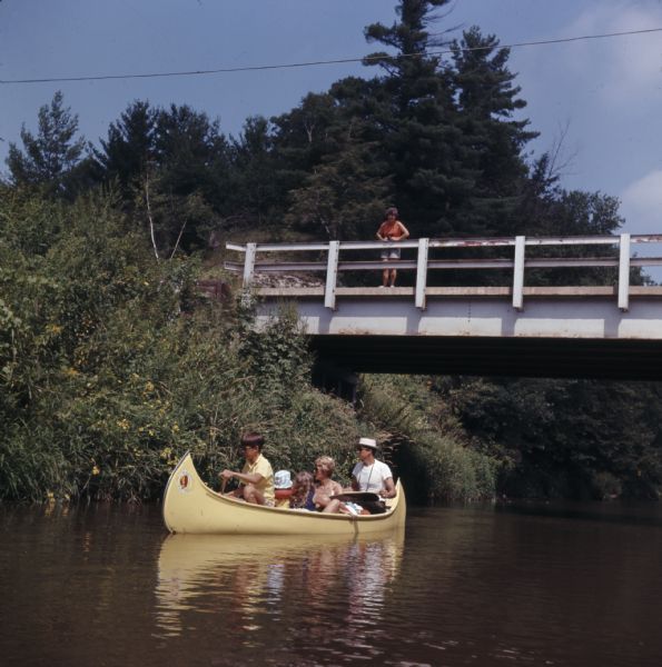 A group of people are sitting in a canoe and paddling down the Kickapoo River. A woman is looking down on them from a bridge.