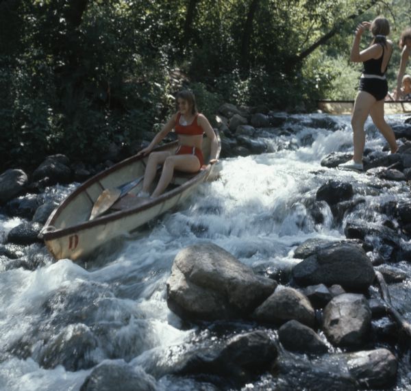 A young woman is riding in a canoe down a rocky rapids on the Crystal River. Another woman is standing on the rocks, as other people are waiting their turn to canoe on the rapids.