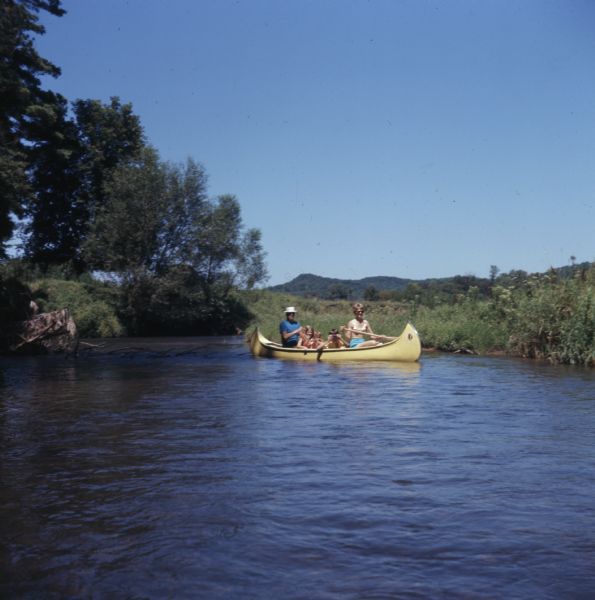 A family canoeing along the Kickapoo River. The mother and father are paddling the canoe carrying three young children sitting in the middle.