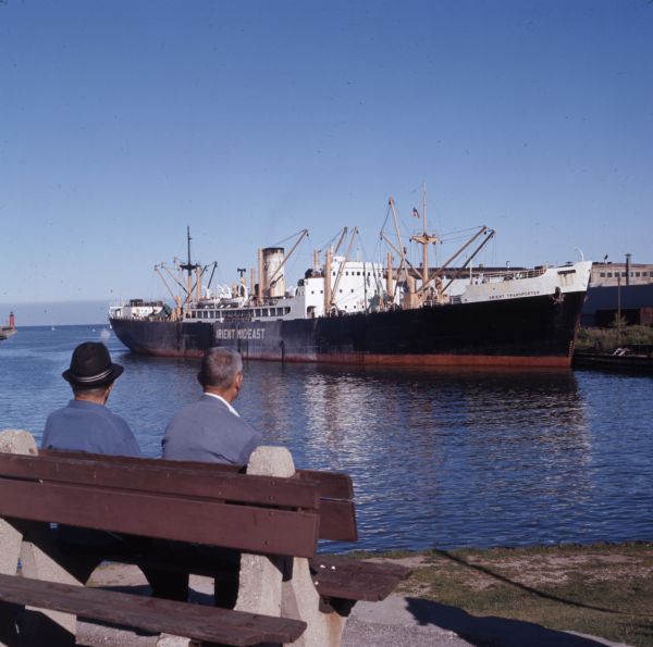 View from behind of two men sitting on a park bench who are looking at the lake steamer docked in the harbor. In the distance, a red lighthouse is at the end of the harbor.