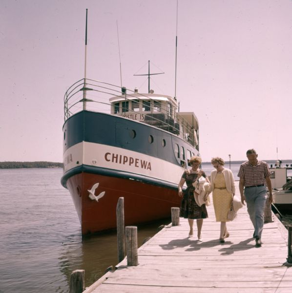 Two women and a man are walking down the dock near a boat named "Chippewa" on Rocky Island.c
 South Twin Island is across the water in the background.
