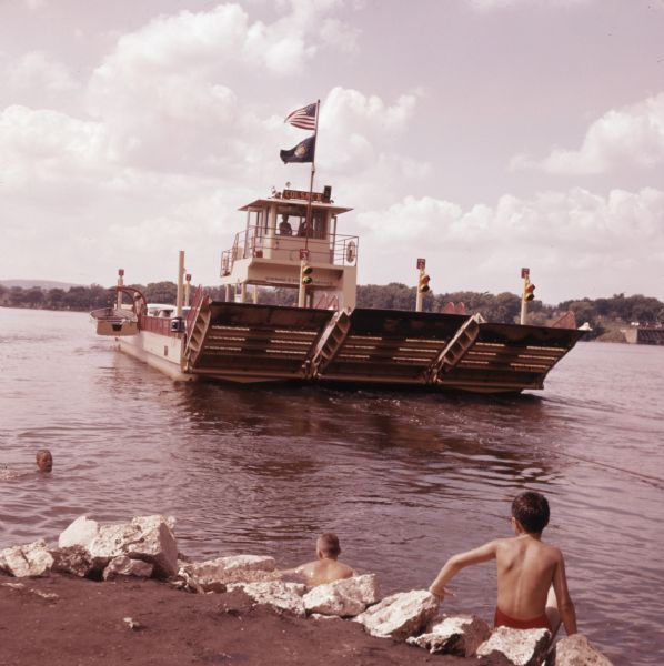 A car ferry moving out across the Wisconsin River. Three boys are swimming in the river in the foreground.