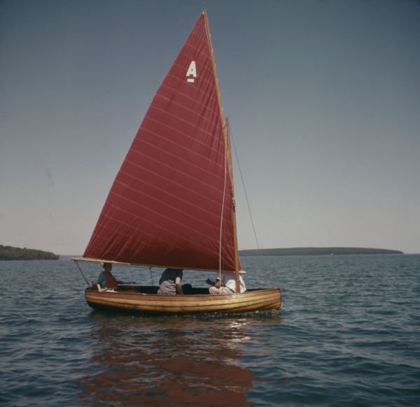 Three people are sitting in a small wooden sailboat with a red sail. Trees are covering the shoreline in the background.