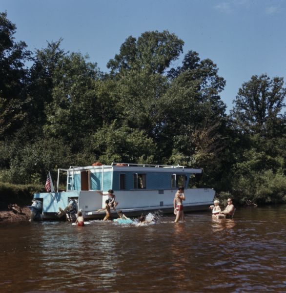 A group of children and adults swimming and playing in the water of the Wolf River next to their blue houseboat docked at the shoreline.