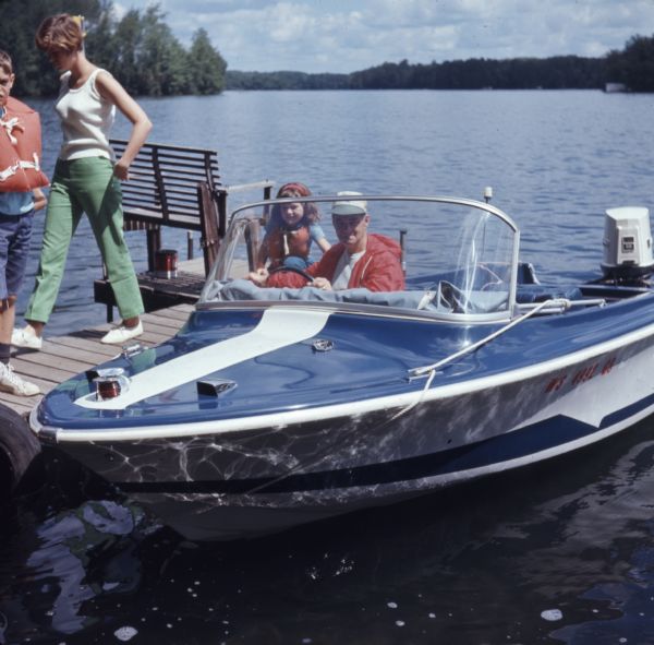 A man is sitting in a motorboat behind the steering wheel. A girl wearing a life jacket is behind him kneeling on the dock. Walking on the dock on the left is a young woman, and a boy on the far left is standing and wearing a life jacket. In the background are trees along the shoreline.