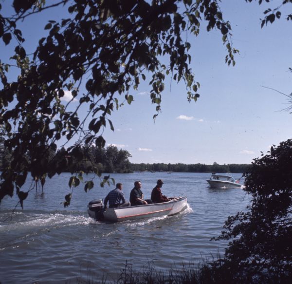 View from shoreline of three men sitting on a small motorboat with their fishing rods on Waupaca Chain O'Lakes. Other boats are on the lake in the distance.