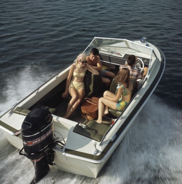 Elevated view of two young men and two young women sitting in a motorboat. The driver is wearing a shirt, and the two women and the other man are wearing bathing suits. A picnic basket is between the seats.