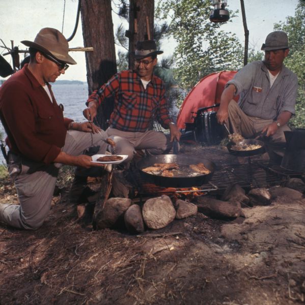 Three men are sitting around a campfire cooking meat and hash on iron skillets. There are trees and a lake in the background.