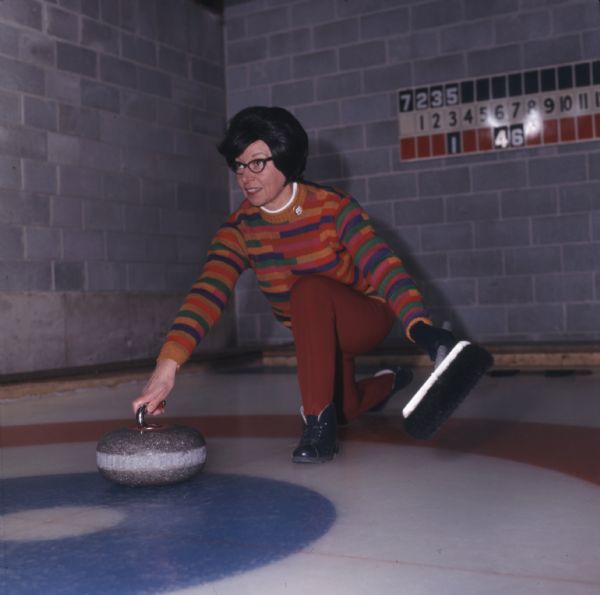 A woman is leaning over the ice preparing to deliver the stone in a curling game. She is holding a broom in her left hand and the stone in her right.